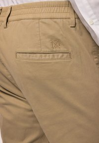 Khaki broek met een platte achterzak en een gestructureerde tailleband. Geborduurd logo-detail in de rechterbovenhoek van de zak.