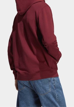 Persona con sudadera con capucha color granate y vaqueros azules de mezclilla, vista desde atrás contra un fondo claro y sencillo.