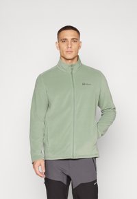 Fleecejack in lichtgroen, volledig met rits en hoge kraag, met zijzakken en een logo op de linkerborst. Zachte textuur.