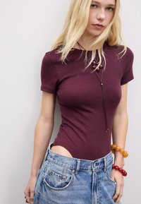 Burgunderfarbene kurzärmlige Body mit strukturierter Oberfläche, kombiniert mit hellblauen, hochgeschnittenen Jeansshorts. Perlenarmbänder setzen farbige Akzente.