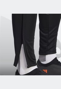 Černé sportovní kalhoty s rozepínatelným větracím otvorem u kotníku, doplněné bílými žebrovanými ponožkami a černými botami s oranžovými akcenty.