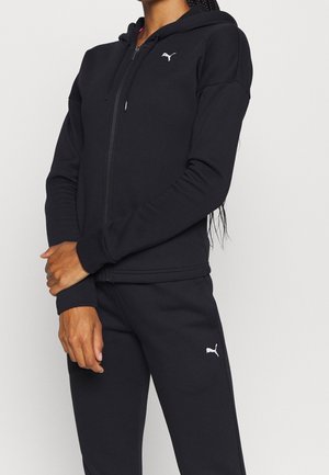 Person trägt schwarzen Puma-Zip-up-Hoodie und passende schwarze Puma-Sweathosen, steht mit verschränkten Armen vorne.