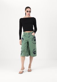 Fekete, hosszú ujjú crop top zöld farmer culotte-tal párosítva, amely tengerészkék batikolt mintákkal díszített. A modellen napszemüveg és bézs szandál van.