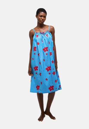 Femme debout pieds nus portant une robe bleue ample avec des rayures verticales blanches et des motifs floraux rouges, fines bretelles et un décolleté froncé.