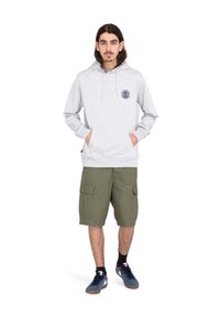 Sudadera gris clara con bolsillo tipo canguro y cordón, que presenta un logo circular en azul marino en el pecho. Llevado con pantalones cortos de cargo oliva y zapatillas.
