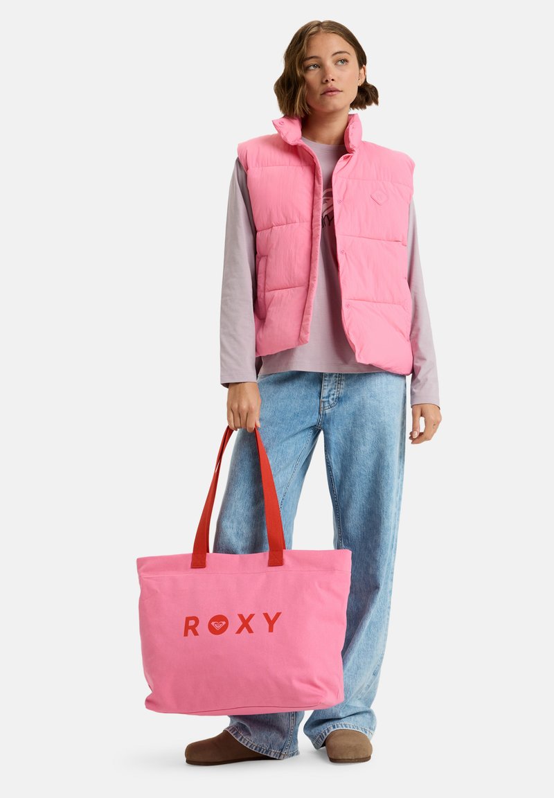 Roze gewatteerde vest met hoge kraag, gedragen over een lichtgrijs shirt, gecombineerd met wijde blauwe jeans en een roze schoudertas met rode "ROXY" tekst.