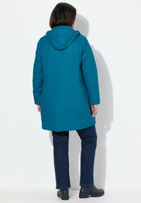 Tealfärgad vattentät jacka med huva, med långa ärmar och rak passform. Kombinerad med mörkblå jeans och svarta ankelstövlar.