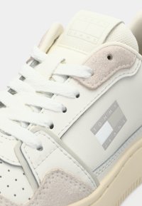 Zbliżenie białego sneakersa Tommy Jeans z beżowymi zamszowymi akcentami, białymi sznurówkami oraz logo po bokach i na języku.