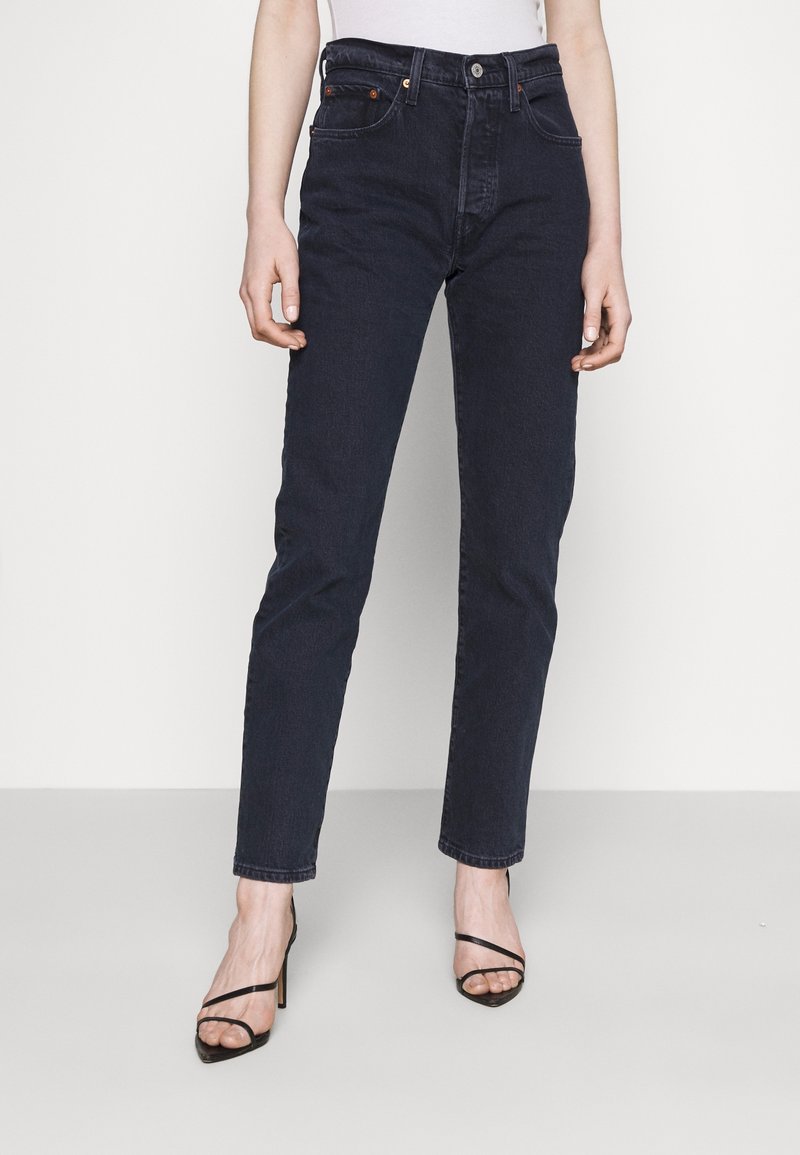 Personne portant un jean taille haute bleu foncé et des sandales à talons hauts noires avec des lanières, debout devant un fond clair uni.