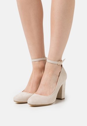 Chaussures à talons hauts en daim beige avec un bout rond, une bride à la cheville et un talon épais, présentant une texture lisse et un design minimaliste.