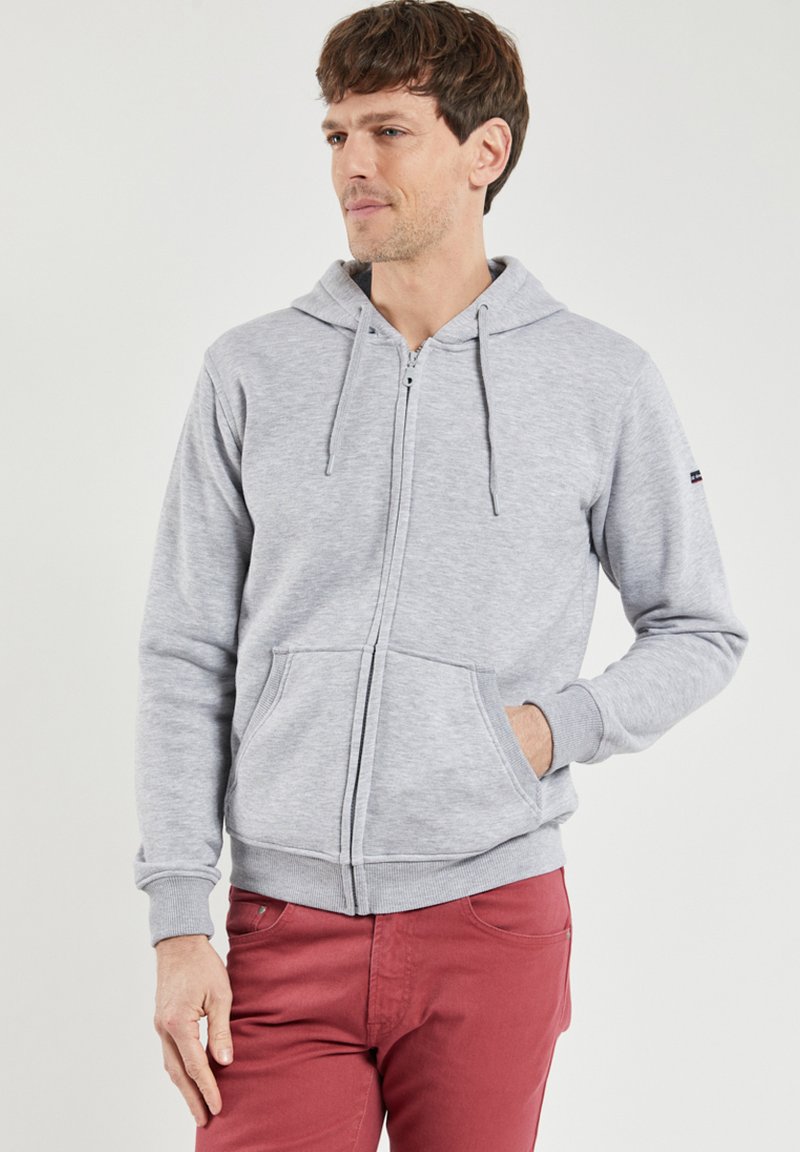 Hoodie zippé gris en tissu doux, avec une poche kangourou à l'avant, un capuchon avec cordon de serrage et des poignets côtelés. Porté avec un pantalon rouge.