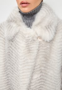 Faux-Fur-Cape mit horizontalen Streifen in sanftem Weiß und hellgrau, einem runden Kragen und einem einzigen Knopfverschluss.