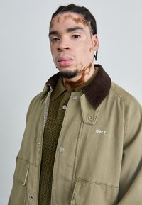 Olijfgroen jack met een bruine corduroy kraag, drukknoopsluitingen en een geborduurd "OBEY"-logo. Textuur gebreid sweater eronder.