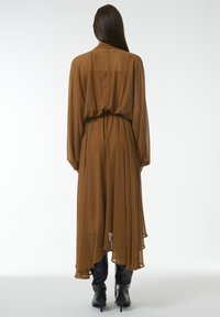 Robe brune fluide et transparente avec des manches longues et une taille cintrée. Présente un ourlet asymétrique et une texture superposée. Portée avec des chaussures noires.