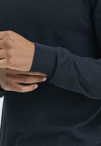 Marineblå genser med ribbestrikket mansjett, laget av et mykt, teksturert materiale. Nærbilde av hender som justerer ermet, som viser et tettsittende design.
