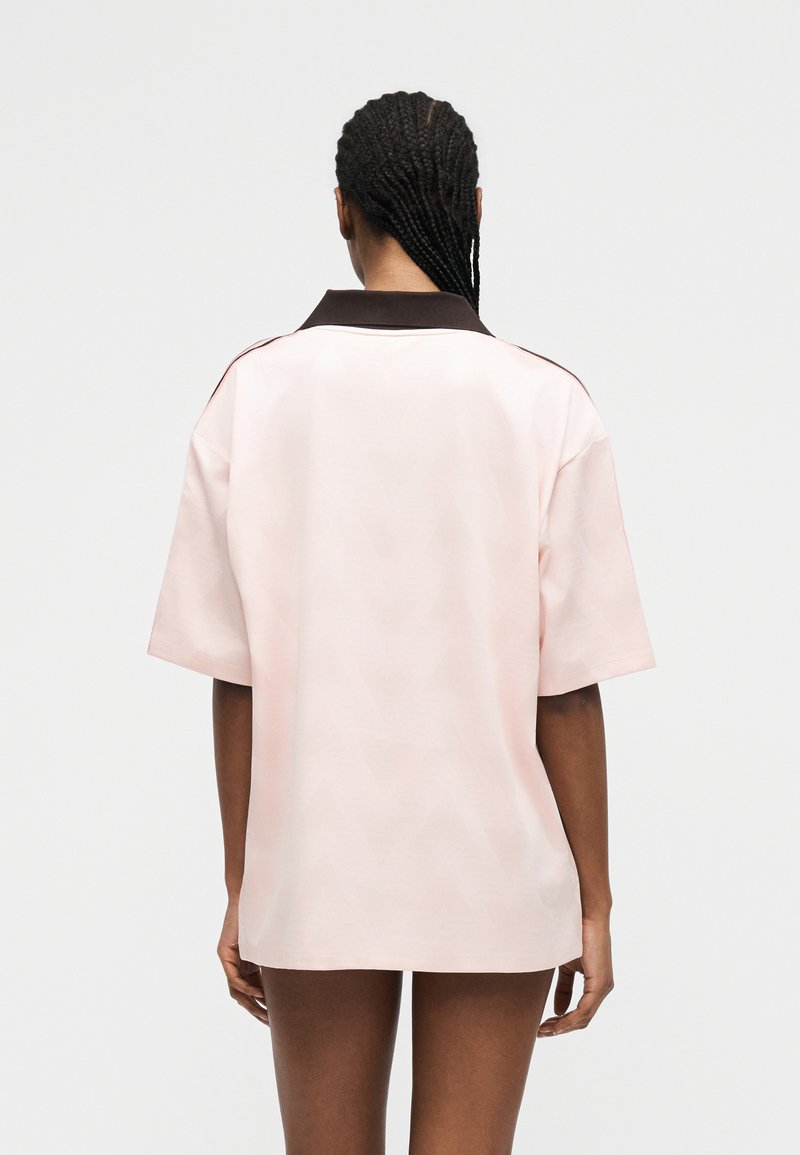 Femme aux cheveux tressés portant une chemise ample rose pâle avec un col foncé, debout de dos devant un fond uni.