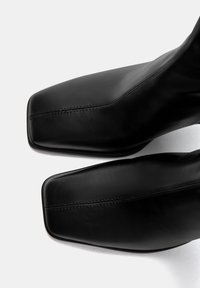 Bottes cheville en cuir noir avec une pointe carrée, un design élégant et des détails de couture minimalistes. Texture lisse et finition brillante.