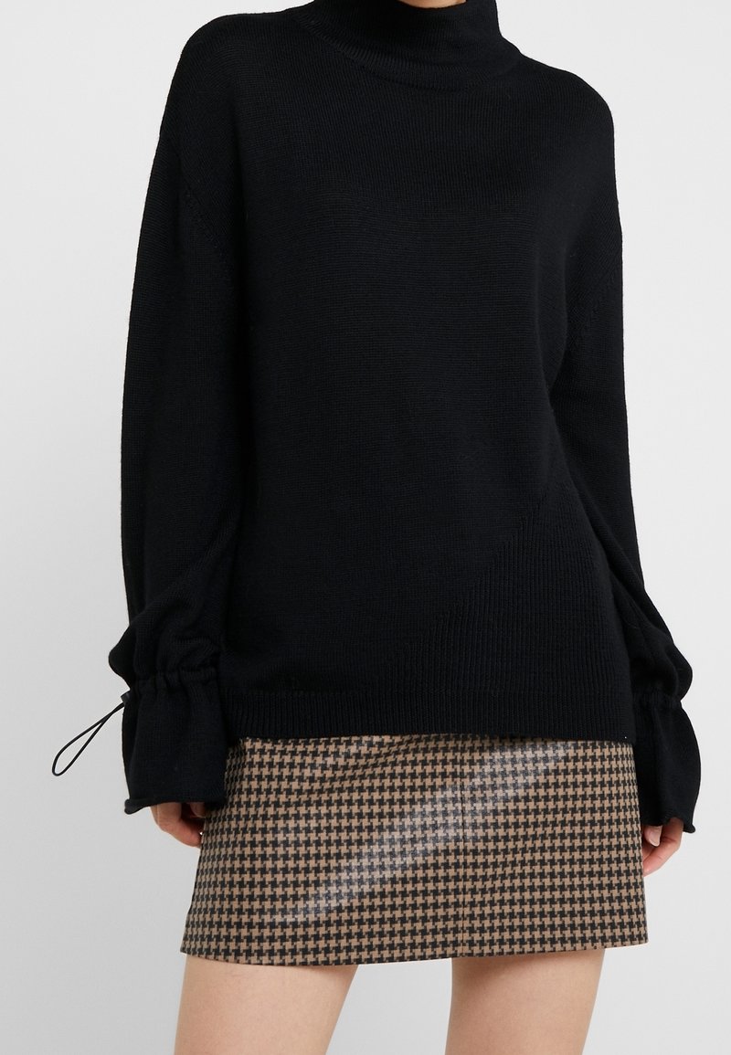 Femme portant un pull noir à manches longues avec des poignets à cordon et une mini-jupe marron et noire à motif pied-de-poule.