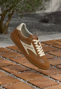 Brun og kremfarget Karl Lagerfeld-sneaker med hvite lisser svevende over trekvadratiske fliser utendørs nær et lite tre.