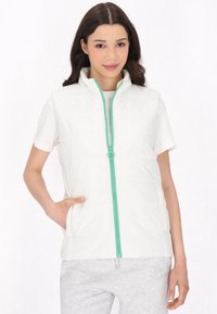 Gilet bianco trapuntato con colletto alto e zip color teal. Abbinato a una camicia bianca a maniche corte e pantaloni felpati grigi.