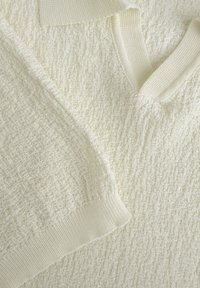 Gelaagd crèmekleurig sweaterstof met een zachte, geribbelde kraag en boorden. Het materiaal lijkt aaibaar met een subtiel weefpatroon en variaties in textuur.