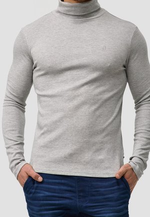 Uomo che indossa un dolcevita aderente grigio chiaro con maniche lunghe e pantaloni blu scuro, con le mani nelle tasche.