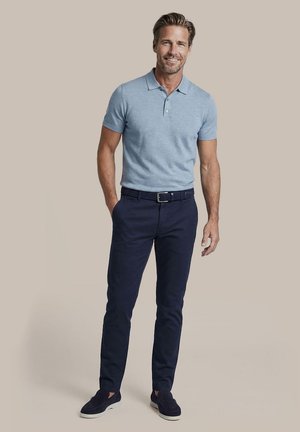 Lachende man die een lichtblauw poloshirt met korte mouwen, donkerblauwe broek met riem en donkere instapschoenen draagt, staand tegen een effen achtergrond.