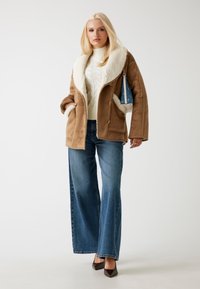 Giacca in shearling marrone con un colletto in pelliccia sintetica crema, abbinata a un maglione bianco a trecce e jeans azzurri a gamba larga. Ballerine nere con punta.