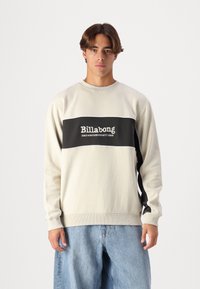 Sudadera de color beige claro con una banda horizontal negra que presenta el texto "Billabong". Puños y dobladillo acanalados, textura suave, corte relajado.
