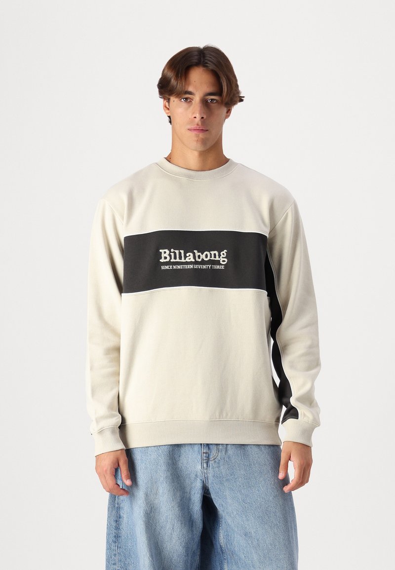 Sudadera de color beige claro con una banda horizontal negra que presenta el texto "Billabong". Puños y dobladillo acanalados, textura suave, corte relajado.