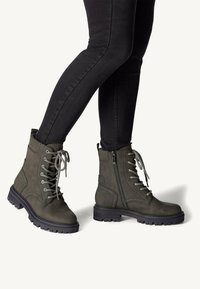 Olive green ankle boots with a textured finish, featuring laces, a side zipper, and a thick black rubber sole with a tread pattern.