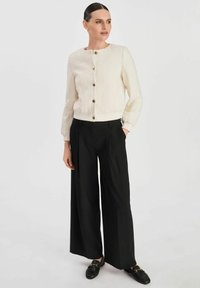 Biała kurtka o teksturowanym wykończeniu z okrągłym dekoltem i złotymi guzikami, w zestawieniu z czarnymi spodniami o szerokich nogawkach i czarnymi loafersami. Gładki materiał z prążkowanymi mankietami.