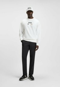 Felpa bianca con logo "BOSS", pantaloni neri e sneaker nere. Il cappellino presenta "BOSS" in nero su uno sfondo bianco.