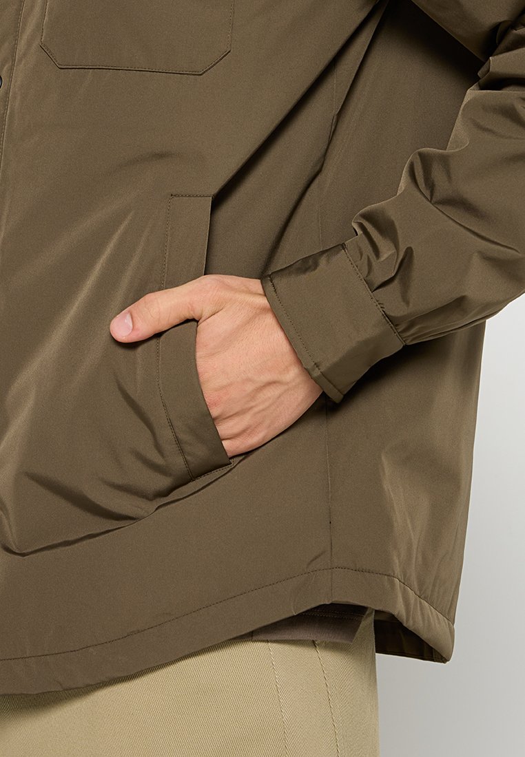 Veste marron, légère, résistante à l'eau, avec une poche avant et des poignets élastiques, présentant une texture lisse et un design décontracté.