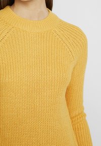 Gelber Strickpullover mit rundem Ausschnitt, Raglanärmeln und strukturiertem Muster. Der Stoff ist weich und leicht dick.