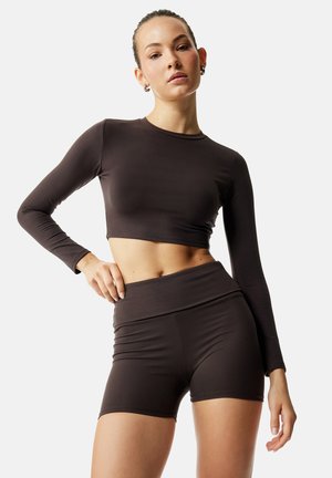 Kobieta ubrana w ciemnobrązowy krótki top z długim rękawem i dopasowane spodenki z wysokim stanem, pozująca z jedną ręką na biodrze na jednolitym tle.
