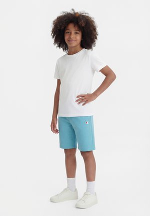 Niño con cabello rizado está de pie con la mano en la cadera, viste camiseta blanca, pantalones cortos azul claro con logo, calcetines blancos y zapatillas blancas sobre fondo blanco.
