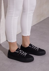 Schwarze Leinenschuhe mit weißen Schnürsenkeln und Gummisohlen, die ein kleines Logo-Tag an der Zunge und eine strukturierte Außensohle aufweisen.