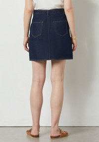 Džínová sukně s přiléhavým pasem a dvěma zadními kapsami. Tmavě modrá barva, klasický střih a čisté linie. Nosená s hnědými nazouvacími sandály.