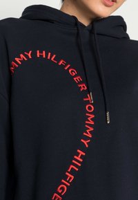 Granatowy hoodie o luźnym kroju, z dużym, czerwonym haftowanym napisem "TOMMY HILFIGER" z przodu oraz sznurkiem ściągającym z złotymi akcentami.