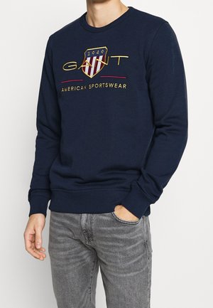Mężczyzna noszący granatową bluzę z haftowanym herbem "GANT 1949" i napisem "American Sportswear", zestawioną z szarymi dżinsami.