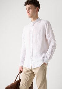 Witte linnen overhemd met lange mouwen en een mandarinenkraag, gecombineerd met beige broek en eenbruine leren tas.