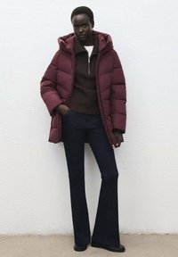 Chaqueta acolchada burdeos con capucha, suéter marrón de canalé, jeans de mezclilla oscuros acampanados y botines negros contra un fondo blanco.