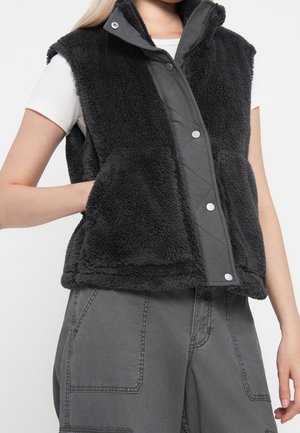 Gilet noir sans manches en matière duveteuse avec boutons-pression, porté sur une chemise blanche à manches courtes, associé à un pantalon gris avec de grandes poches à l’avant.
