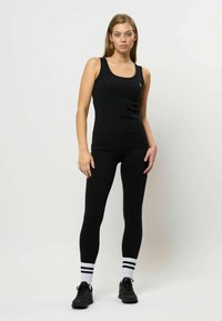 Črna rebrasta majica s tankimi naramnicami v kombinaciji s črnimi legicami. Bele športne nogavice imajo črne črte. Model stoji na nevtralni podlagi.