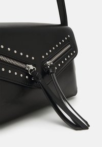 Róg czarnej skórzanej torby z srebrnymi ćwiekami, dwoma srebrnymi suwakami i długimi skórzanymi uchwytami suwaków na białym tle.