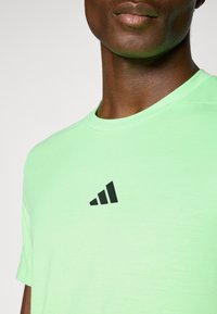 Lichtgroen sportshirt gemaakt van gladde stof; voorzien van een zwart logo met drie strepen op de borst, een ronde halslijn en korte mouwen.