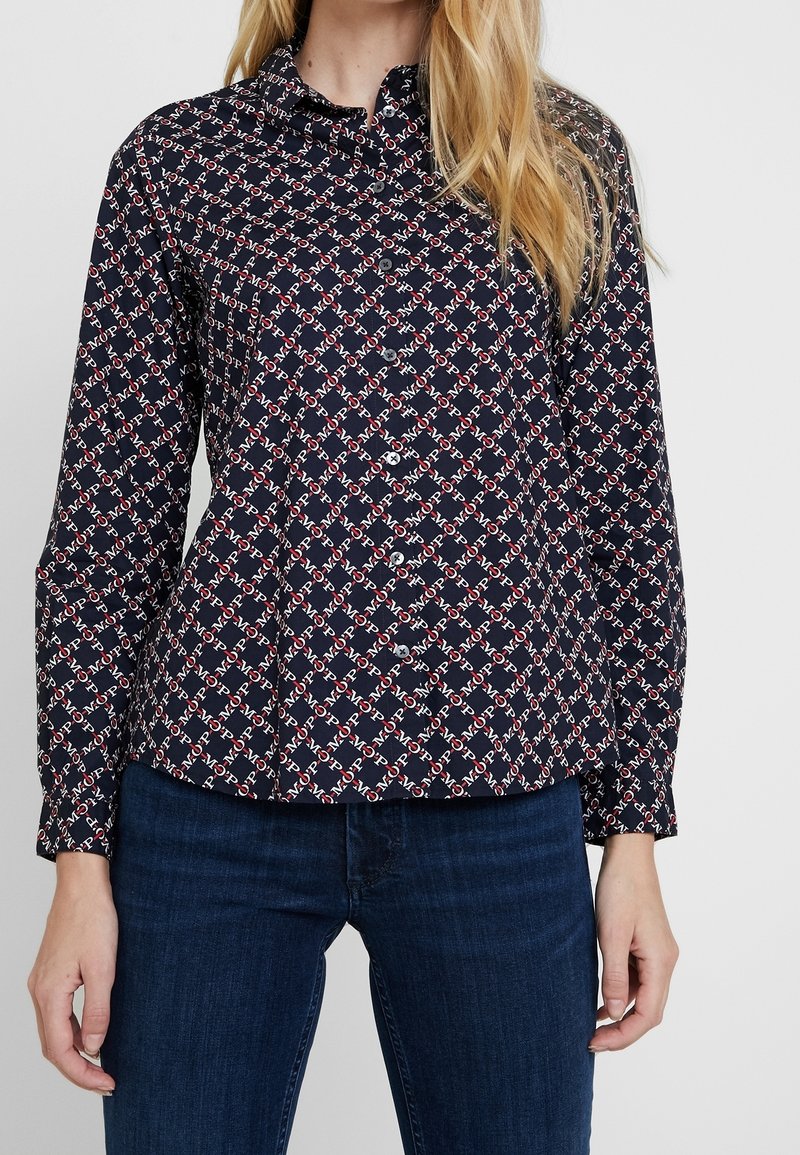 Camicia blu navy con bottoni, caratterizzata da un motivo geometrico in rosso e bianco. Maniche lunghe e collo, realizzata in un tessuto liscio.