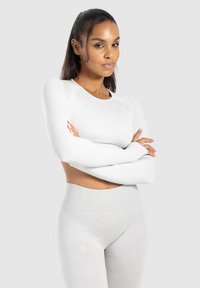 Zelfverzekerde vrouw met lang donker haar in een witte crop top met lange mouwen en hoge taille leggings, staande met gekruiste armen op een grijze achtergrond.