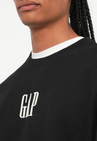 Svart hettegenser med rund halsringning og hvit krage, teksturert stoff, og brodert "GAP"-logo i hvitt på brystet.